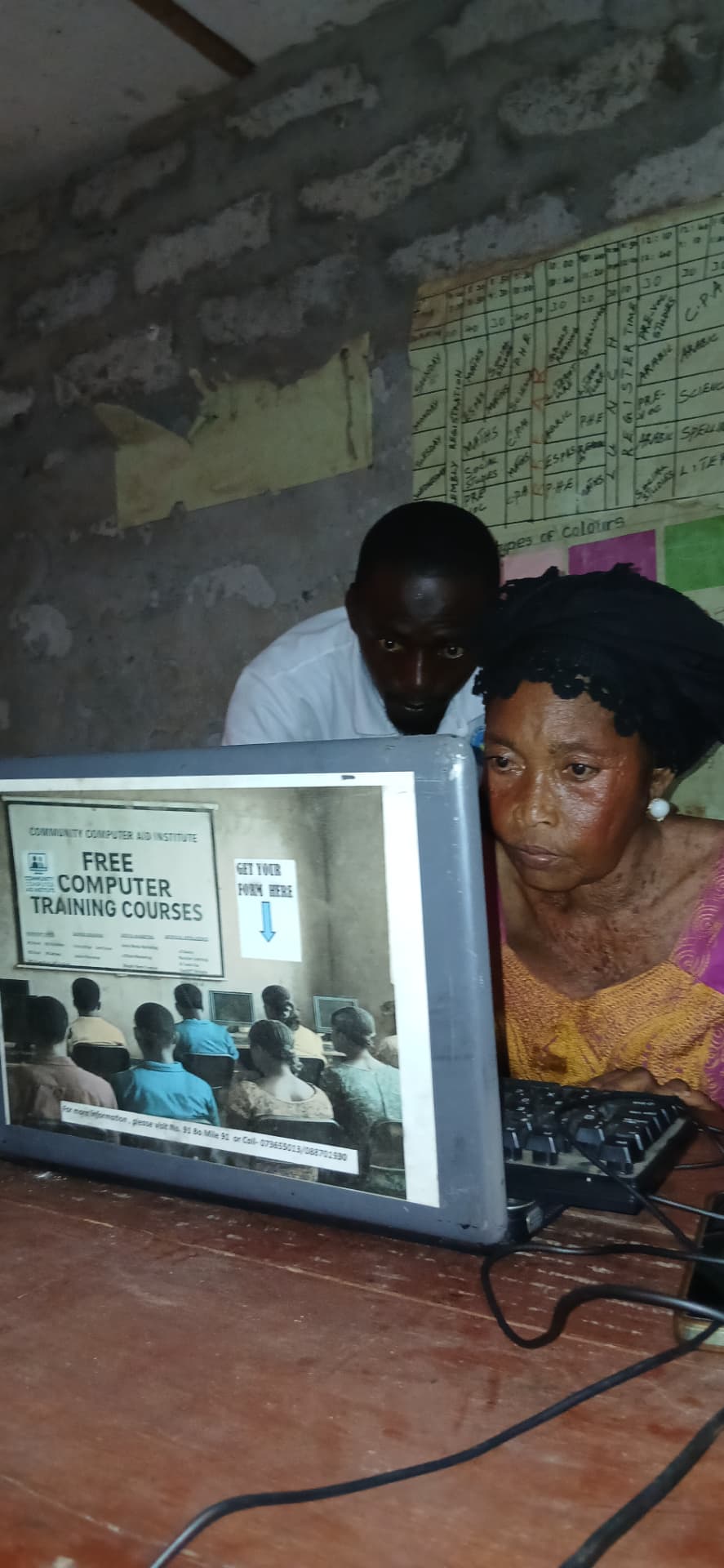 An older woman learning on a desktop computer with a trainer beside her and a Free Computer Training Courses poster on the screen