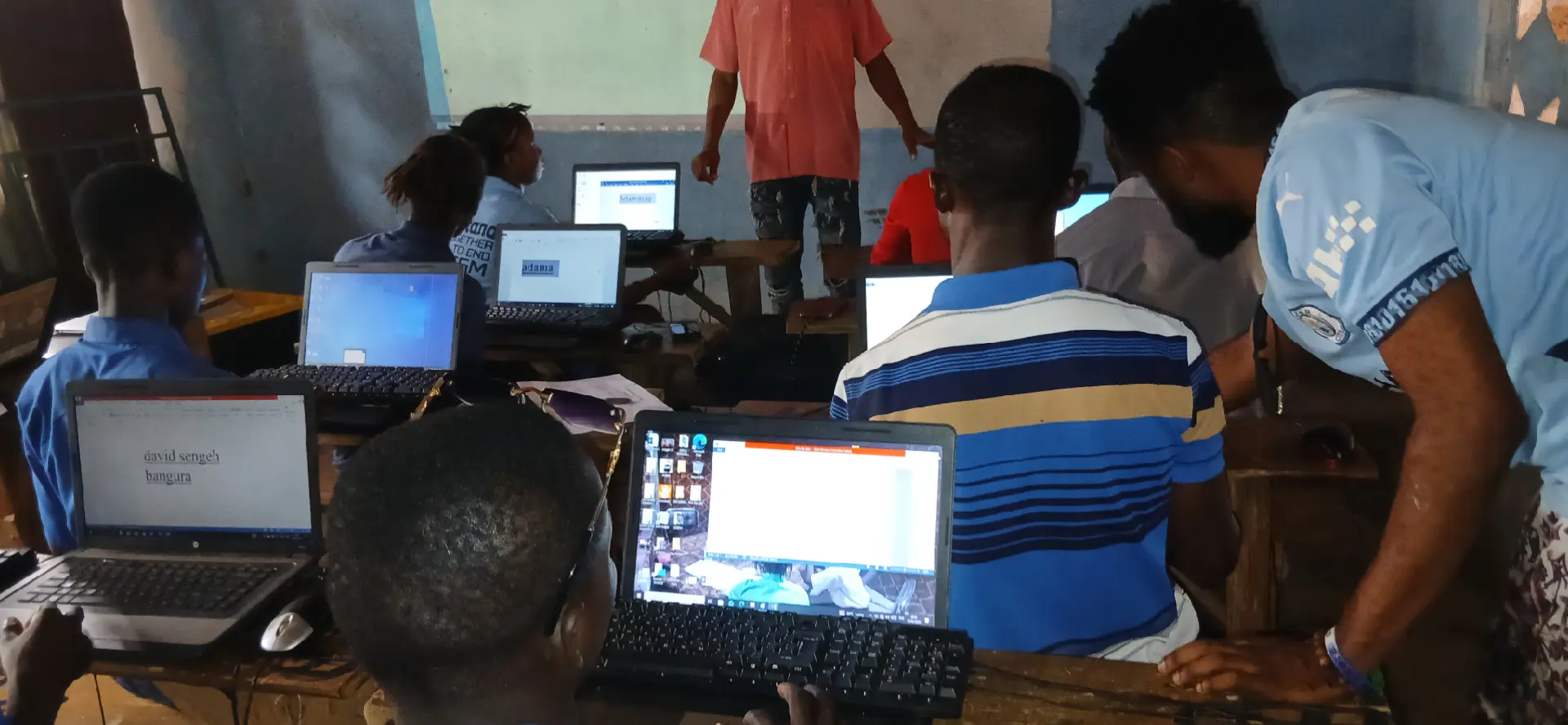 A full classroom of learners typing on laptops with a facilitator assisting
