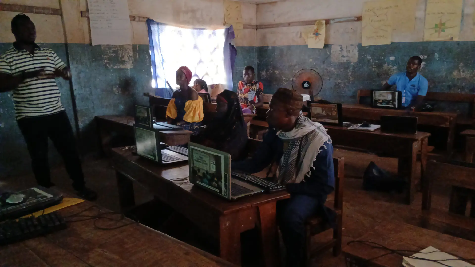 Women and men of mixed ages learning on laptops in a rural classroom with a facilitator standing