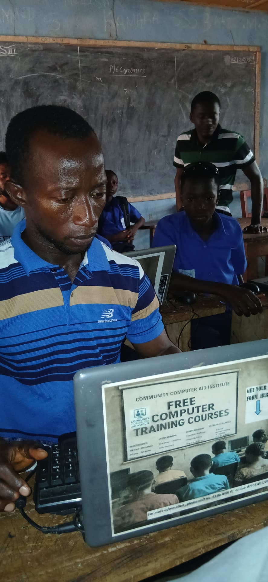 An adult learner practising on a laptop beside pupils, with a CCAP free computer training poster on the screen lid